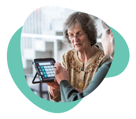 Older woman using an AAC tablet with support from another person, selecting words on the screen together.