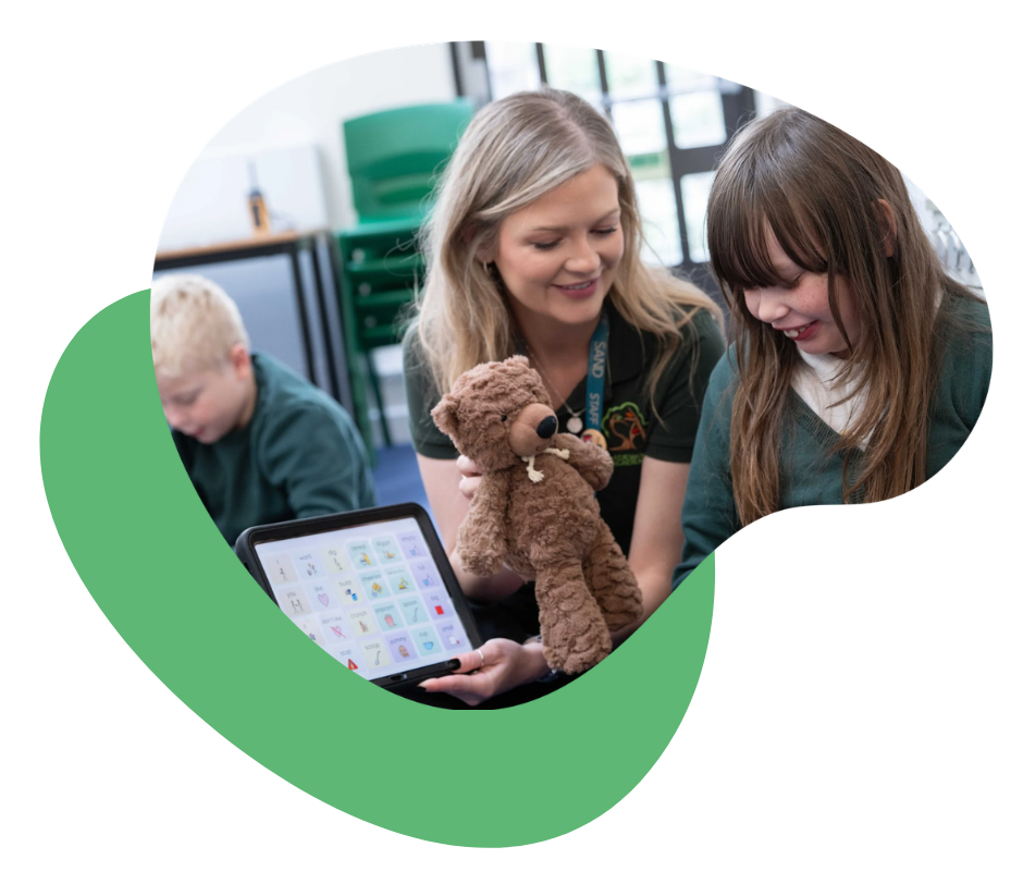 Adult modelling to a child using an AAC app on a tablet while playing with a teddy bear in a classroom setting.
