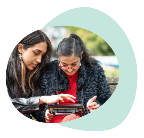 Two people sit together outdoors using an AAC device. One person points to symbols on the screen while the other looks on