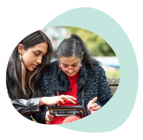 An AAC user sitting next to her sister on a bench, using an AAC device together.